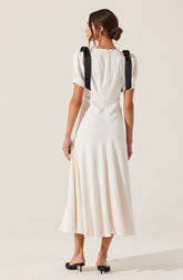 A woman is wearing an elegant, white dress that features a fitted bodice and a flowing, pleated skirt reaching mid-calf. The dress has short, puffed sleeves and black ribbon shoulder details tied into bows. She is also wearing black, open-toe, low-heeled shoes.